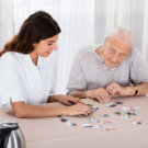 Elder Woman Playing Jigsaw Puzzle On Table With Her Nurse