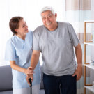 Female Caretaker Assisting Happy Senior Man While Walking At Home